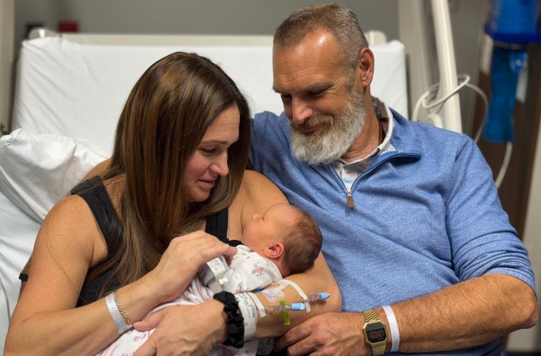 A mother and father sit closely together in a hospital bed, lovingly cradling their newborn baby. The mother has medical bands on her wrist, and both parents are smiling warmly as they gaze at their child.