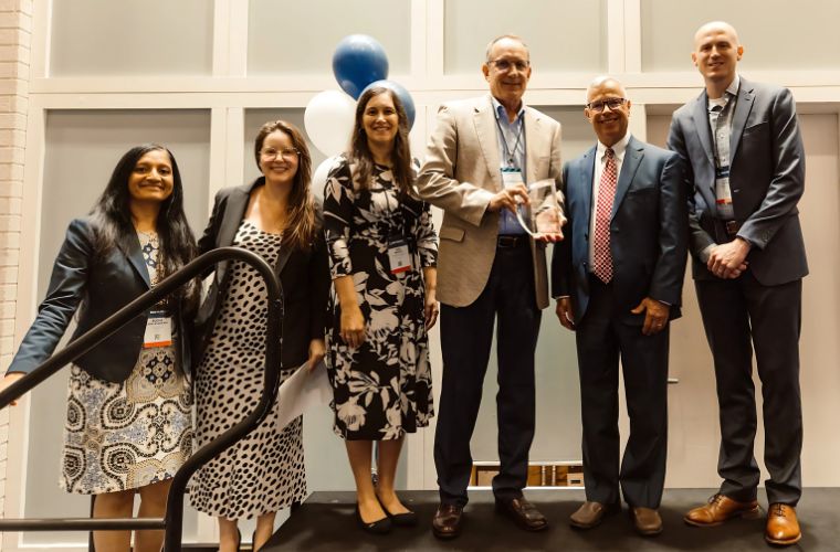 A group of six professionally dressed individuals stands on a stage smiling. The man in the center holds a clear award plaque, with blue and white balloons in the background, suggesting a celebratory event or recognition ceremony.