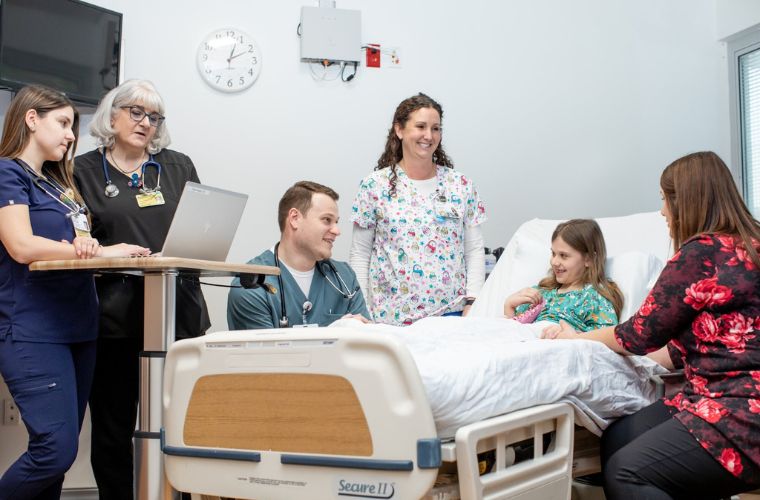 A group of healthcare professionals, including a nurse standing with a laptop and a doctor, interact with a pediatric patient in a hospital bed and a visitor seated beside the bed. The room appears well-equipped and everyone is engaged in a cheerful conversation.