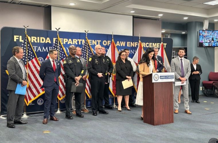 A group of individuals, including law enforcement officers and civilians, standing at a press conference. One person is speaking at a podium with a sign reading "Department of Law Enforcement." The background displays several American flags and the podium bears the seal of Florida Attorney General James Uthmeier.
