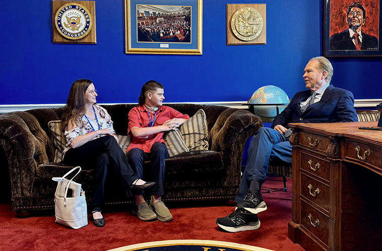 A woman and teen boy sit on a couch talking to a man in a chair in a room with blue walls and red carpet.