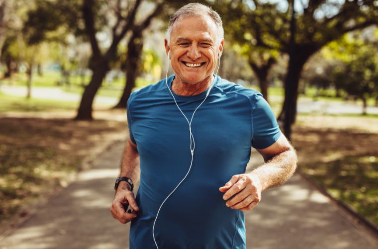 An older man wearing athletic clothing and headphones, jogging in a park surrounded by greenery and trees.