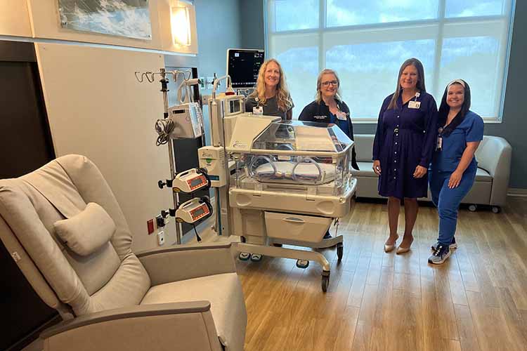 Four medical professionals standing in a hospital room with medical equipment including an incubator and a recliner.