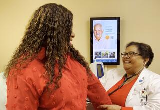 A woman in a red shirt engages in conversation with a nurse practitioner in a clinical setting.