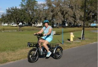 A woman wearing blue shorts and top with a matching blue bike helmet smiles as she rides her electric bike down the road.