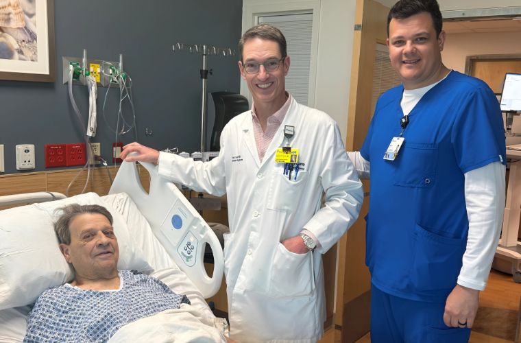 Physician in a white lab coat and nurse in blue scrubs stand next to a patient in a hospital bed.