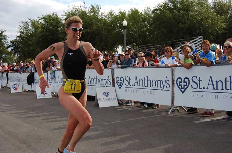 A woman dressed in a swimsuit runs past a crowd of people and signs that advertise a race.