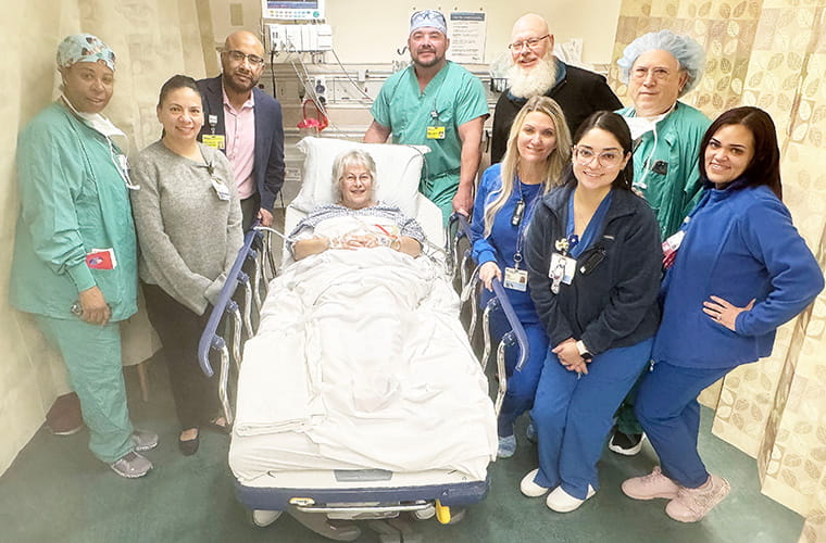 A woman wearing a hospital gown lies in a hospital bed and is surrounded by a group of people, some dressed in hospital scrubs and others in regular clothes.