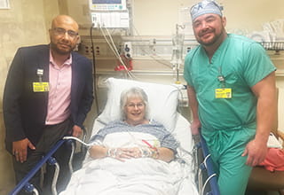 A woman lying in a hospital bed is flanked by two men, one dressed in a jacket and the other in hospital scrubs. 