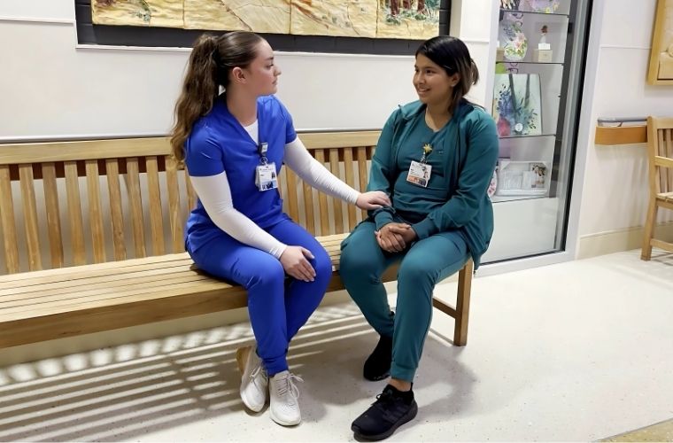 Two women sit on a bench inside a hospital corridor. The woman on the left is wearing blue scrubs and holding the arm of the woman on the right, who is wearing green scrubs.