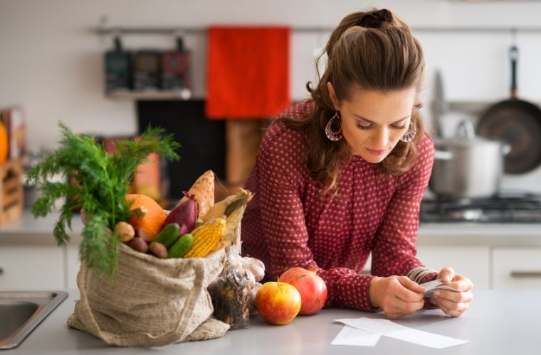 A young woman is glancing down at receipts from a grocery shopping trip in her kitchen. Sitting on the counter beside her is a canvas bag filled with various fruits and vegetables.