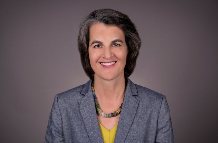 A smiling headshot of Keri Eisenbeis. She is wearing a gray jacket with a yellow V neck shirt underneath and a statement necklace made up of large, cylindrical beads in a range of earthy tones. She has short, straight graying hair.  