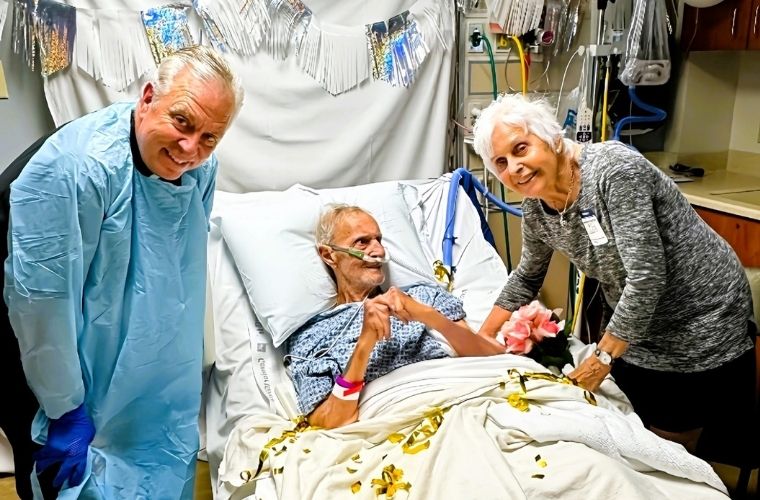 A hospital patient, Anthony, is lying in a bed with white sheets decorated with gold ribbon pieces, wearing a patterned gown and a pink wristband. The patient is holding hands with a visitor, Nancy, standing on the right, who is leaning slightly forward, smiling and holding a bouquet of pink flowers. Another person on the left, Father John, is wearing a full blue protective gown and gloves, standing close to the bedside. Behind the bed, silver and white fringe garlands are draped as festive decorations. The background includes medical equipment, tubing, and cabinets, with a white balloon visible near the upper right corner.