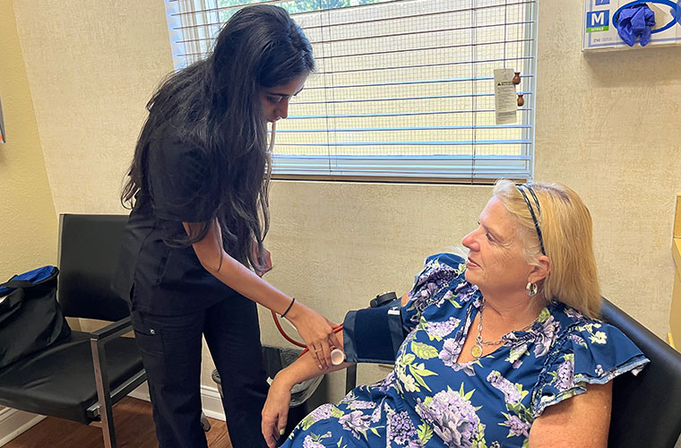 A woman with long dark hair wearing black medical scrubs uses a stethoscope and blood pressure cuff to take the blood pressure of a woman with blond hair wearing a flower print dress who is sitting in a chair at a doctor's office.