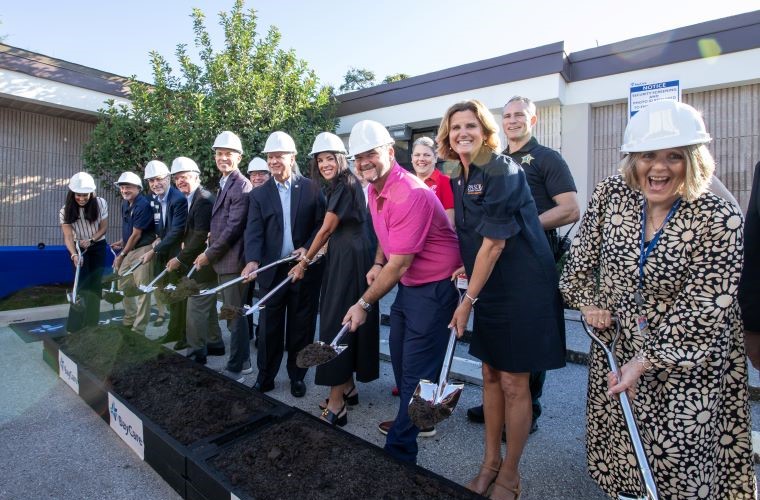 Ten people hold shovels over a sandbox at a groundbreaking event.