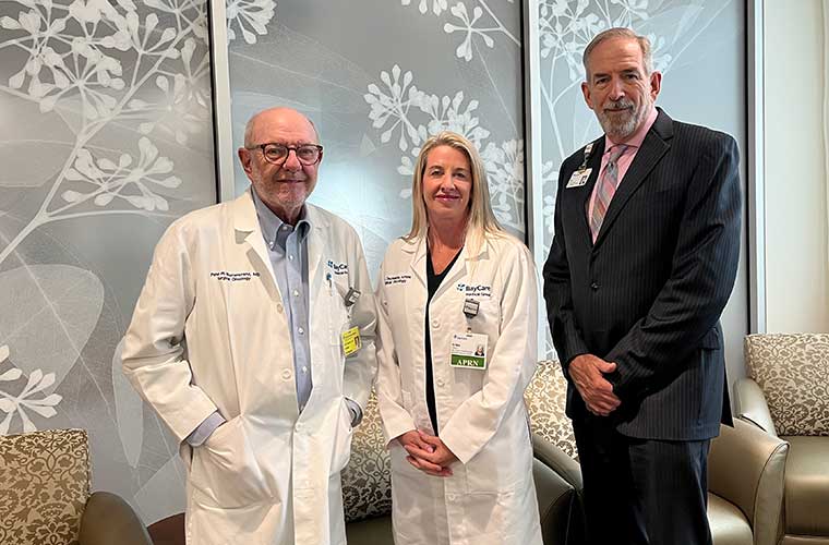Three people -- a man and woman wearing medical coats and another man in a business suit -- stand in a lobby area in front of some windows with a design printed on them.
