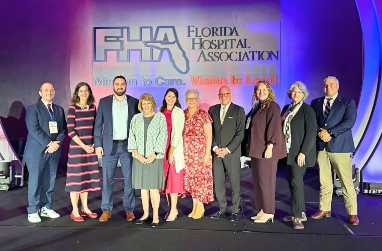 A group of nine individuals standing on a stage in front of a large backdrop displaying the Florida Hospital Association logo and the slogan ‘Mission to Care. Vision to Lead.’ The stage is lit with purple and blue lighting, and the group is dressed in professional attire, including suits, dresses, and business outfits.