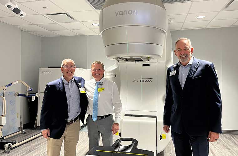 Three men dressed in business clothes stand next to a large piece of medical equipment in a large room.