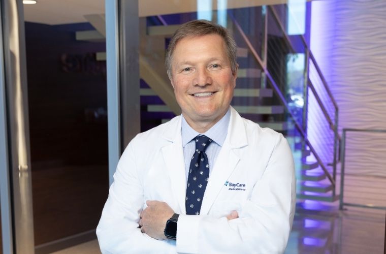 A man smiles as he poses while wearing a white doctor’s jacket over a light blue shirt and a dark blue tie with sailboats on it.