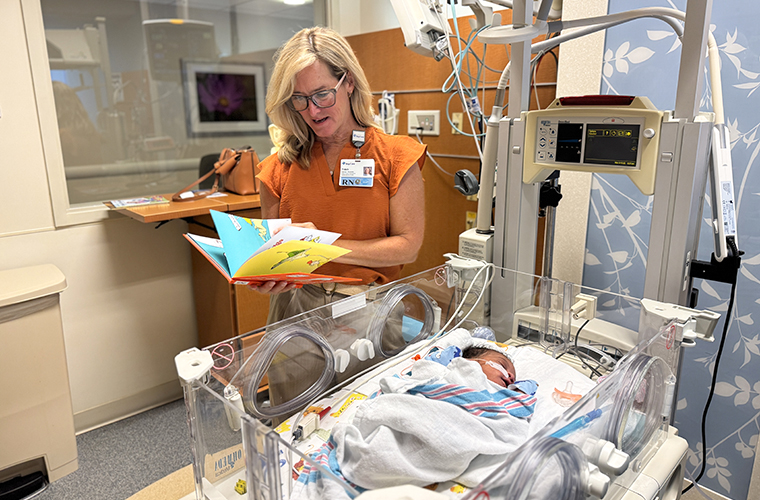 A woman standing next to an infant hospital bed reads a book to a baby. 