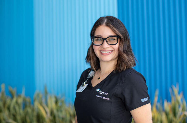 Young woman in black BayCare scrubs wearing square black glasses smiling in front of a blue BayCare building.