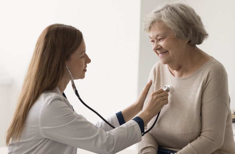 A female doctor with long brown hair listens to an older female patient’s heartbeat with a stethoscope during an exam.