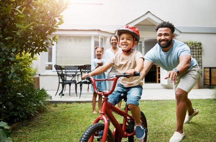 A smiling child wearing a helmet rides a bicycle across a grassy backyard while a parent runs alongside, with two family members watching and cheering near a home.