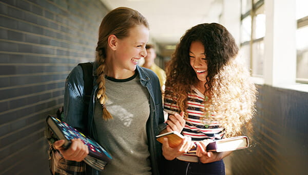 Two middle school aged girls walking together down a hallway, smiling and holding textbooks