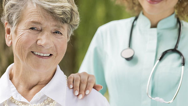 A female nurse places her hand on the shoulder of a smiling senior female patient.