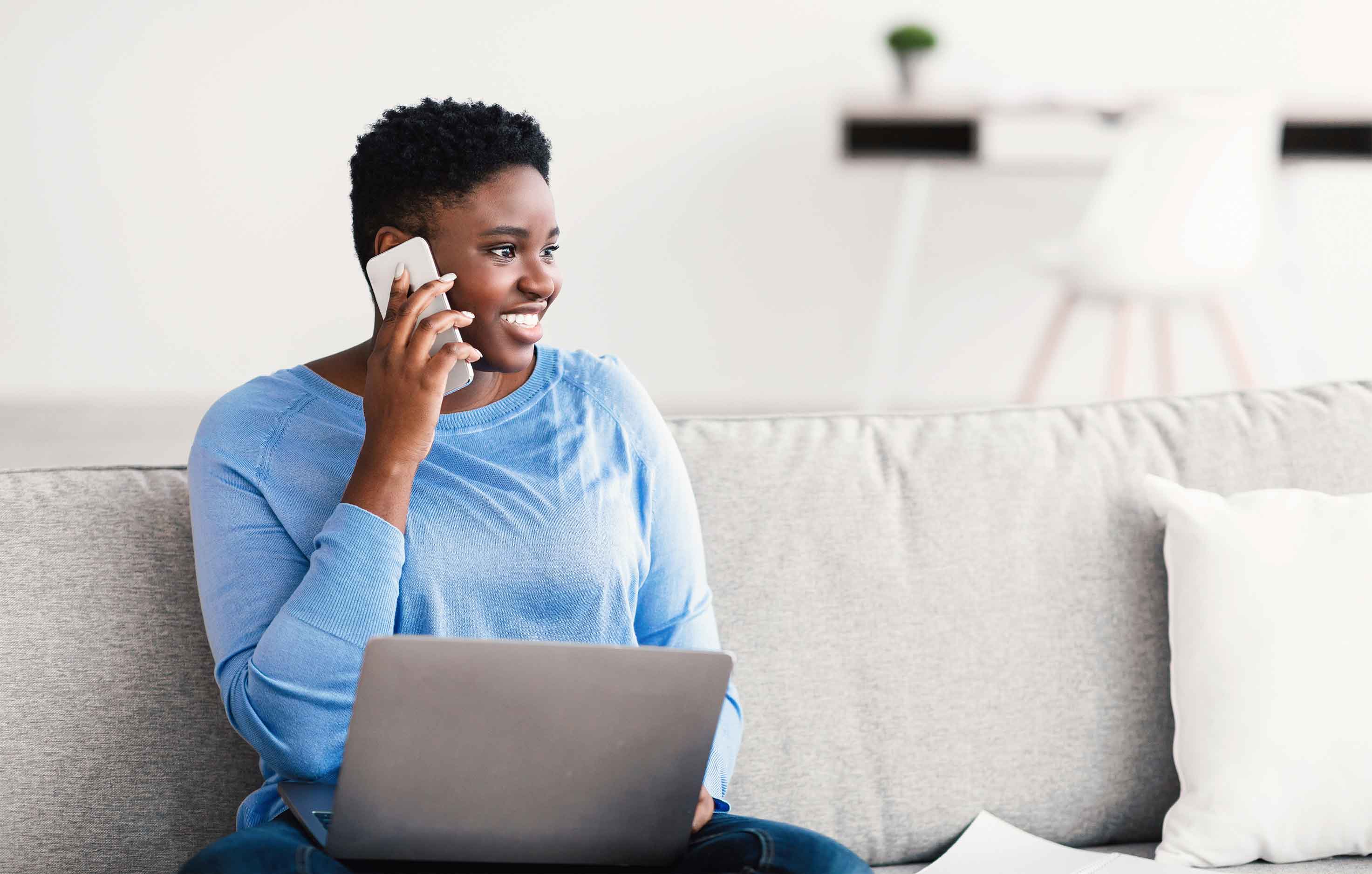 A black female sitting on the couch with a laptop on her lap, making a phone call and smiling in a bright room