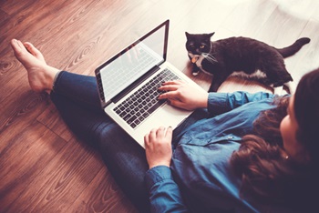 Barefoot woman sitting on floor works on laptop while a cat watches her