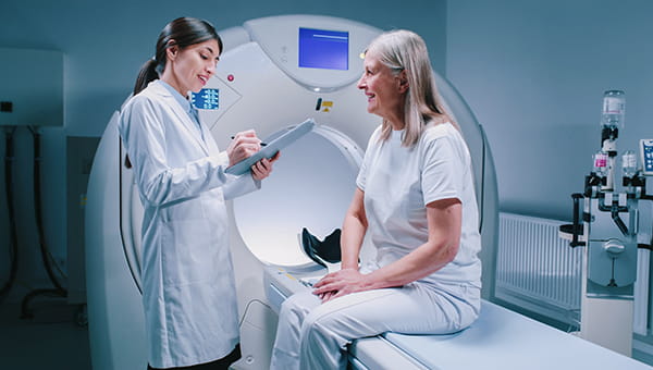 a young female doctor charting next to an elderly female patient on a MRI machine
