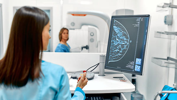 A mammographer looks at an image of breast tissue with the mammogram patient in the background.
