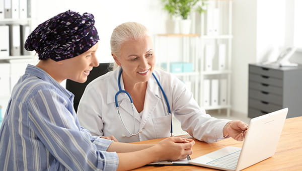 A cancer patient with a head covering looks at a laptop screen with a doctor.
