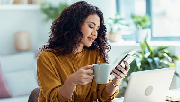 Lady looking at her phone while drinking coffee in front of her laptop