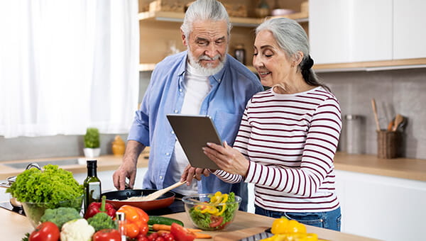 An elderly couple looks at a tablet while cooking in the kitchen.