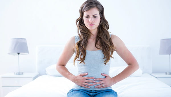A woman is sitting on a bed and holding her abdomen with both hands.