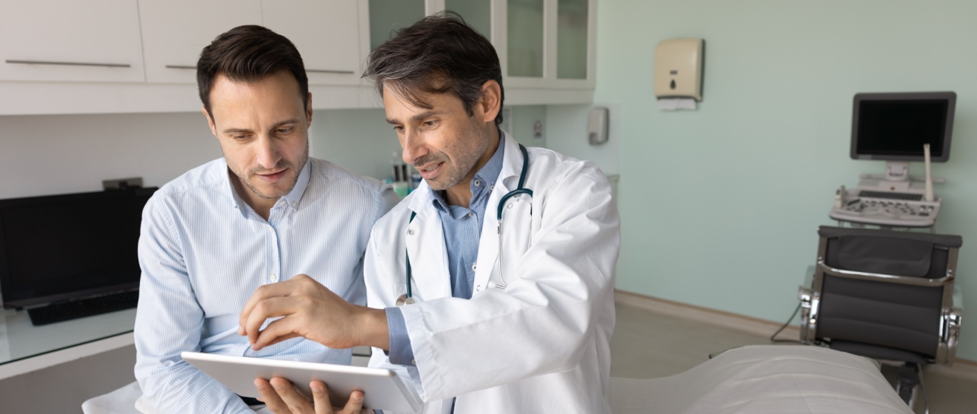White male primary care physician sitting next to a middle aged white male patient while showing an ipad to him