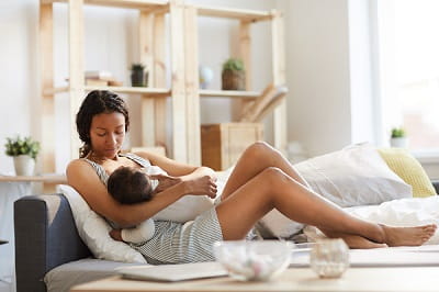 Serious calm careful young black mom sitting on sofa bed and breastfeeding baby while holding son in