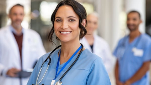 A female nurse smiles with a group of doctors blurred behind her.