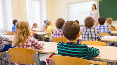 A group of children is sitting in an elementary school classroom listening to their teacher.