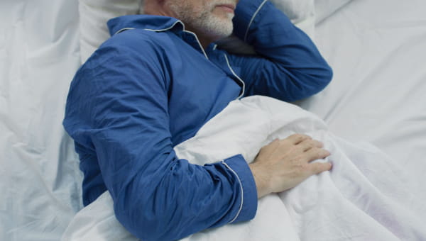 A senior male laying in bed comfortably asleep in blue pajamas.
