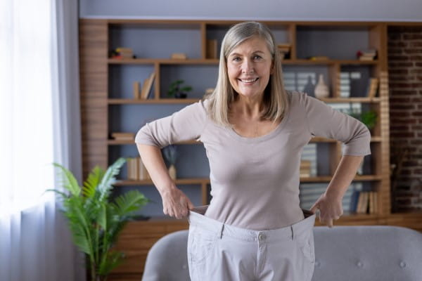 Joyful mature woman stands confidently showing weight loss wearing oversized pants, expressing happiness and success. Her smiling face reflects pride in personal health and fitness accomplishment.